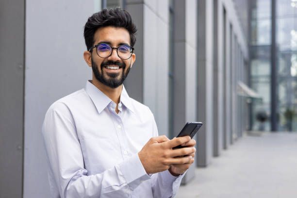 Smiling man in glasses holds a smartphone while standing outside a modern building.
